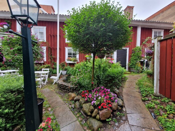 Mysig trädgård med blomster, stenstig, träd, gröngräs och rött trähus, utomhusmöbler, lykta, sommardag.