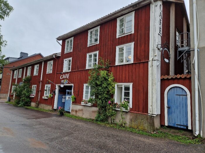 Röd träbyggnad med caféskylt, blå dörrar, fönster med blomlådor, omgiven av vegetation, på en gråmulen dag.