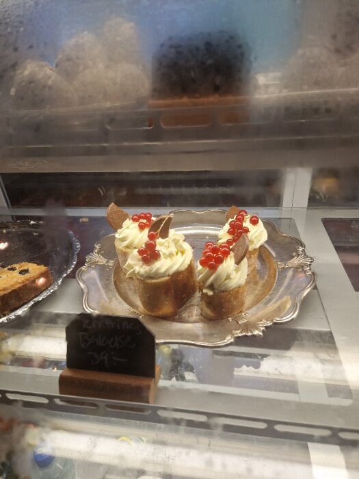 Bakverk i monter, gräddbakelser med bär, prisskylt, reflektion av person, konditori.
