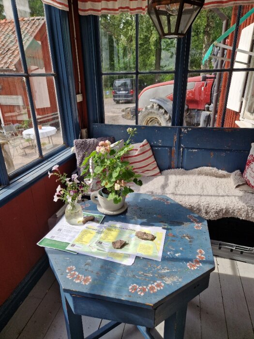 Ett mysigt verandautrymme med blommor, informationsblad på ett bord, utsikt till en trädgård och traktor.