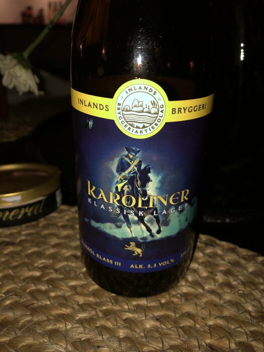 Ölflaska på vävd underlägg, etikett med häst och ryttare, "Karoliner klassisk lager", alkoholhalt 5,3%.