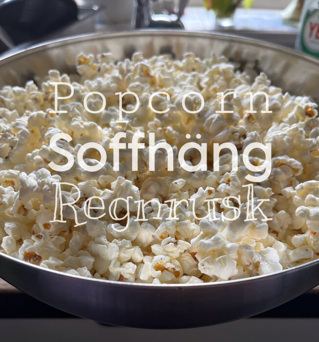 En skål med popcorn, text överlägger: "Popcorn", "Soffhäng", "Regnrusk". Hemmiljö, fönsterljus.