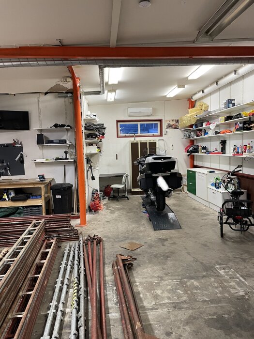 Verkstadsinteriör med motorcykel, hyllor, verktyg och byggmaterial. Arbetskläder hängande. Rött och vitt tema. Rör på golvet.