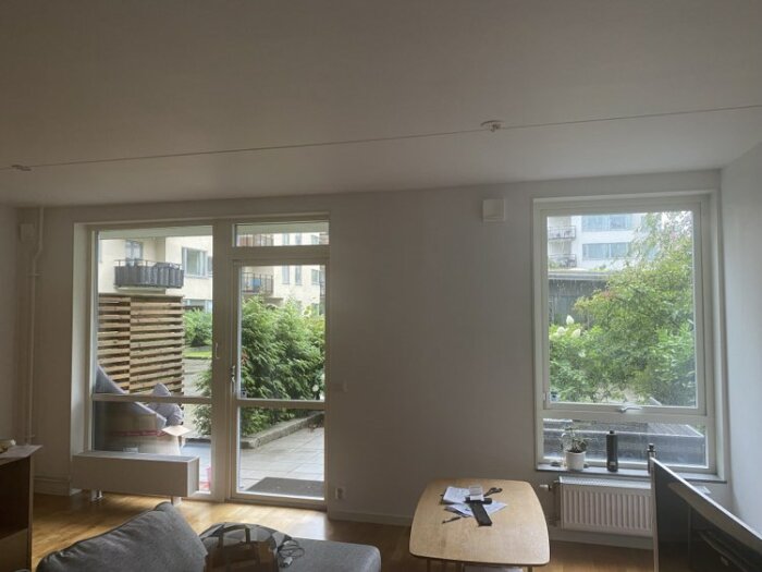 Modernt vardagsrum med glasdörr till uteplats, soffa, bord, fönster med trädutsikt och minimalistisk inredning.