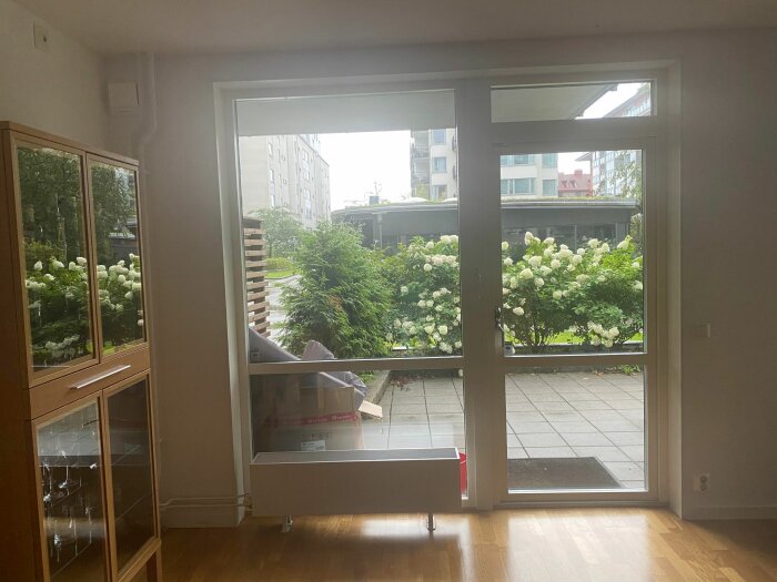Rum med fönsterdörrar öppna mot trädgård, vita blommor, stadsmiljö i bakgrunden, trägolv, möbler.