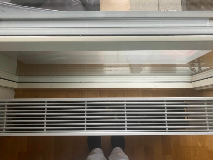 Spår av ett dubbelfönster med radiator och trägolv sett uppifrån, någons skodon i bildens nedre del.