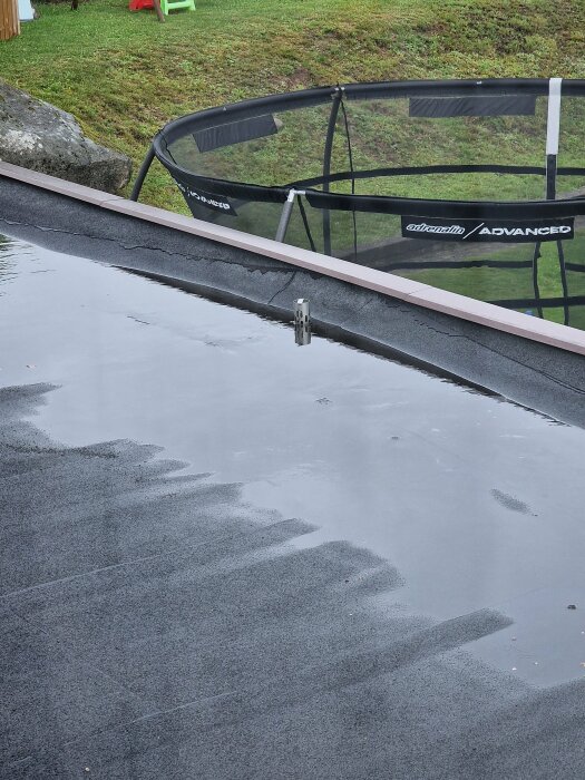 En regnvåt trampolin bredvid en pöl av vatten på en grå trädgårdsterrass.