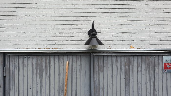 Vit tegelvägg med svart lampa ovanför grå träliknade garageport och en sopkvast.