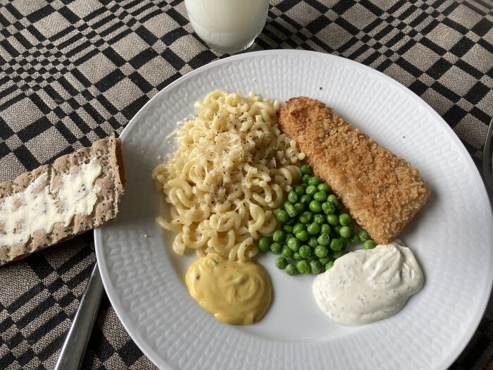 En tallrik mat: pasta, panerad fisk, gröna ärtor, bröd med smör, såser, mjölk. Rutig duk. Hemlagad måltid.