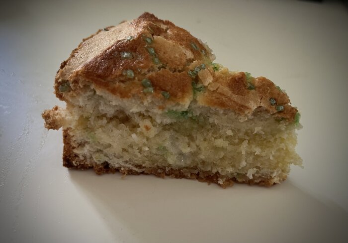 Bit av saftig sockerkaka med grön färgton, strösslad med något grönt, presenterad på ljus yta.