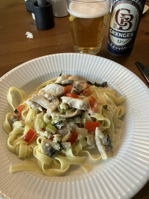 Pasta med kyckling, grönsaker och sås. Öl bredvid. Matservering på vit tallrik. Måltid med dryck.