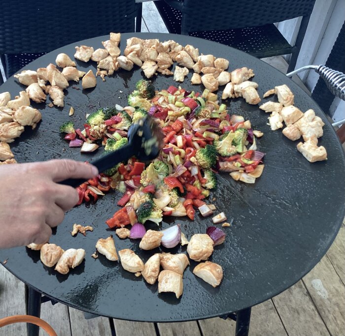 Matlagning utomhus på en stor, rund grill: kyckling, broccoli, paprika och lök. Person vänder grönsaker med spatel.