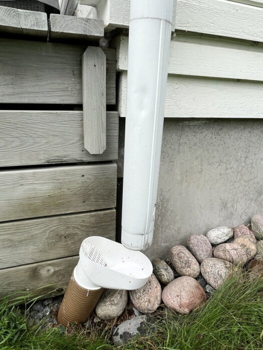 Vitmålat hus, stuprör, mur, vitt avlopp i plast, beigea plastsugrör, stenar och gräs.