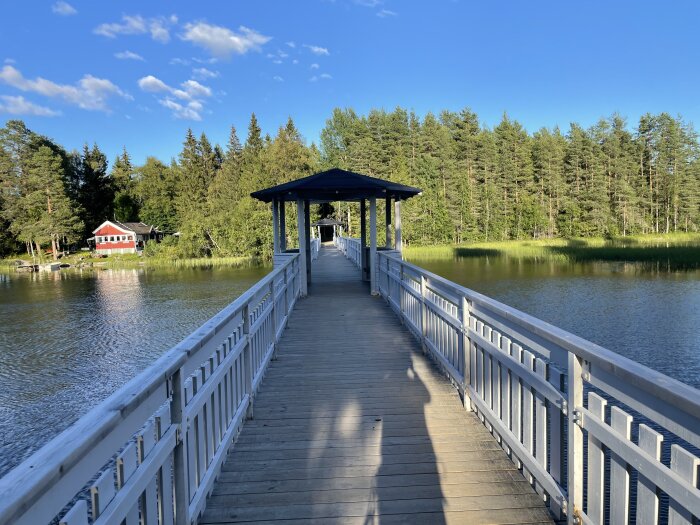 Vit träbrygga leder över en sjö, mot skogskant med klart blå himmel ovanför. Soligt och lugnt landskap.