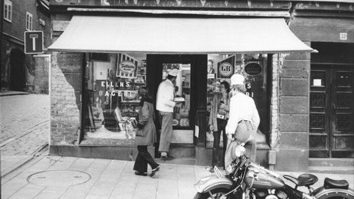 Svartvit bild av personer utanför traditionell bageributik, motorcykel parkerad framför, gatusten, nostalgisk känsla.