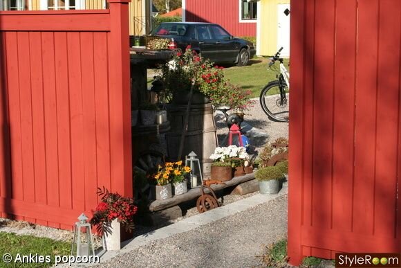 Trädgård med blommor, röda grindar, cykel, antika föremål, och en parkerad bil i bakgrunden.