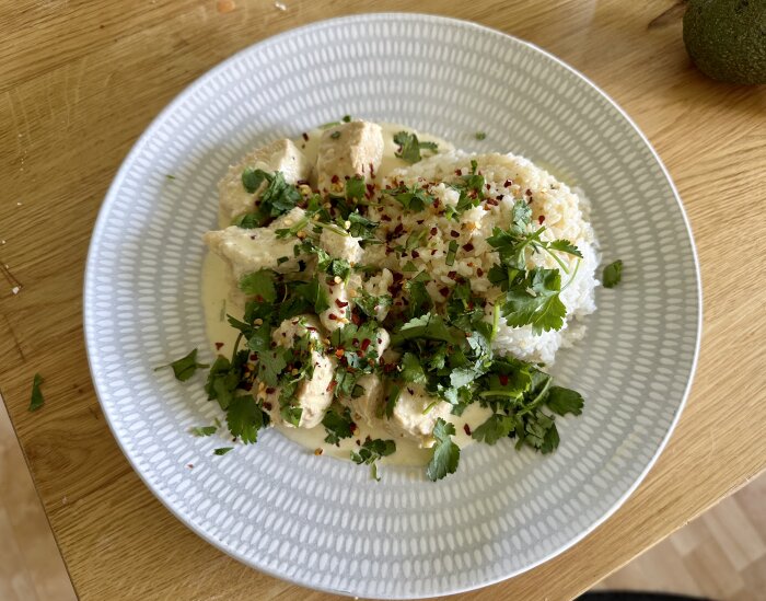 En tallrik med ris och kyckling i sås, garnerad med koriander och chili. Avokado i bakgrunden.