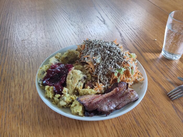 Tallrik med varierad mat, sallad, bacon, solrosfrön, lingonsylt, vattenglas, trämönstrat bord.