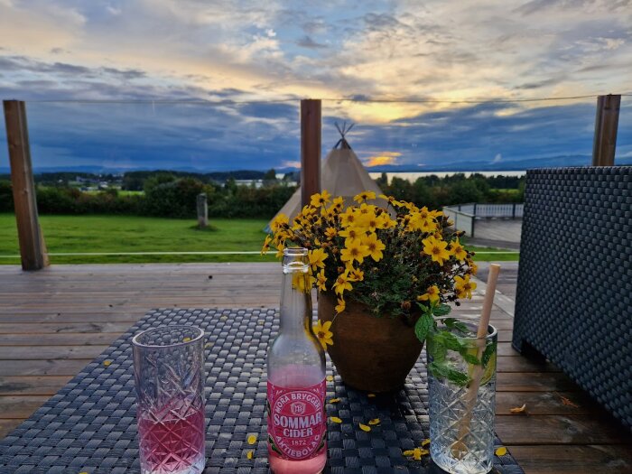 Terrass med utsikt, blomkruka, ciderflaska och glas, avkopplande sommarkvällsstämning.