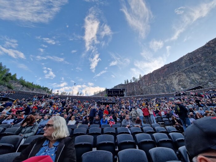 En utomhusarena med publik, blå himmel, klippvägg i bakgrunden, väntande på evenemang.