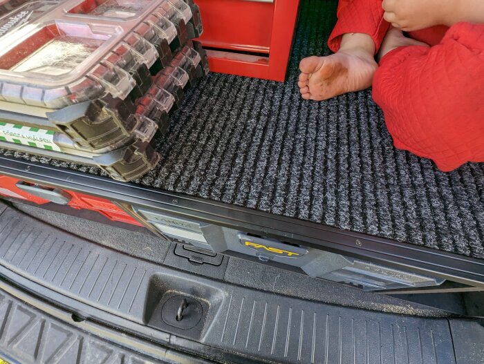 Barns fötter i bilens bagageutrymme bredvid verktygslåda och äggkartonger på en matta.