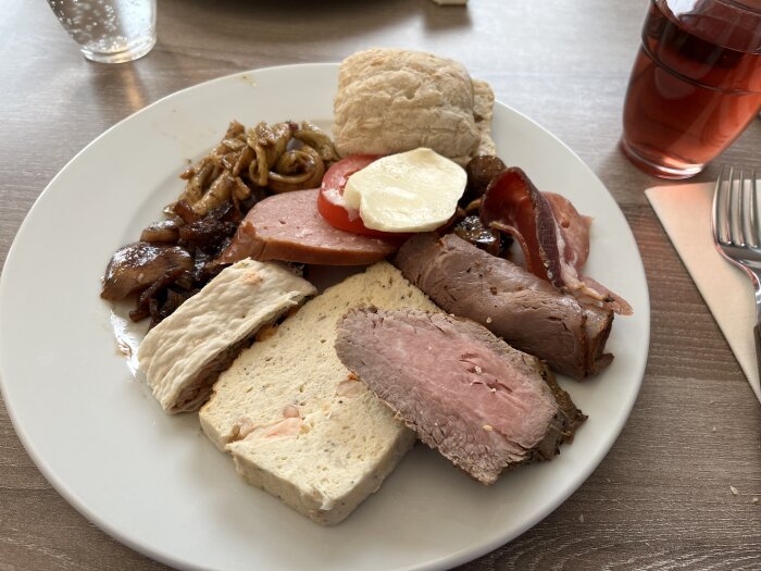 Flera köttbitar, bröd, stekt lök och tomat på en vit tallrik, röd dryck i bakgrunden.