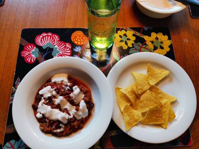 Chili med gräddfil, nachochips, dryck i grönt glas, färgglad duk, träbord.