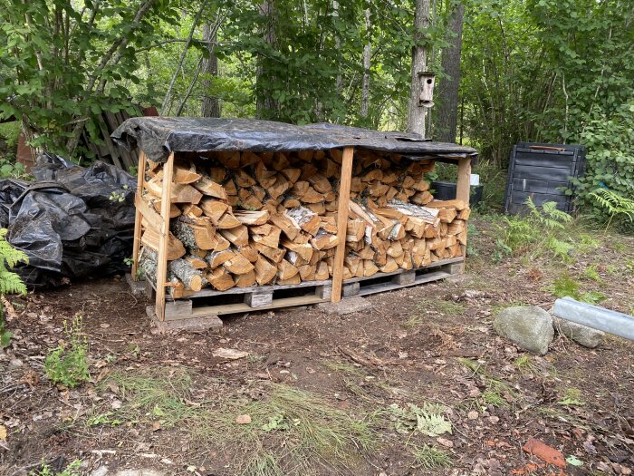 Vedskjul med staplad ved, presenning, skogsbakgrund, kompostbehållare, utomhusmiljö.