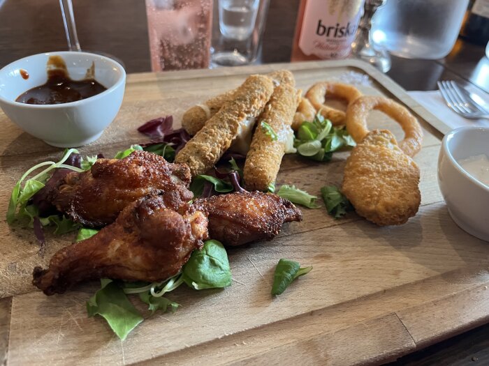 Appetizing assorted fried snacks on wooden board with dips and drink. Chicken wings, mozzarella sticks, onion rings, fresh garnish.