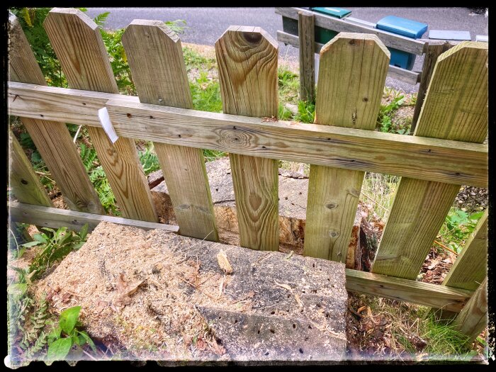 Trästaket med brutna spjälor och trasigt stöd, på en trottoarkant, med grönska och bakgrund med soptunnor.