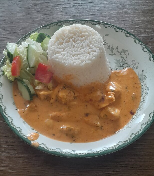 En tallrik med ris, krämig kycklingcurry och fräsch grönsallad serveras på ett mönstrat porslin.