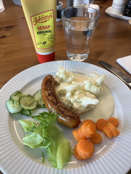En måltid med korv, potatissallad, grönsaker, senap och ett glas vatten.