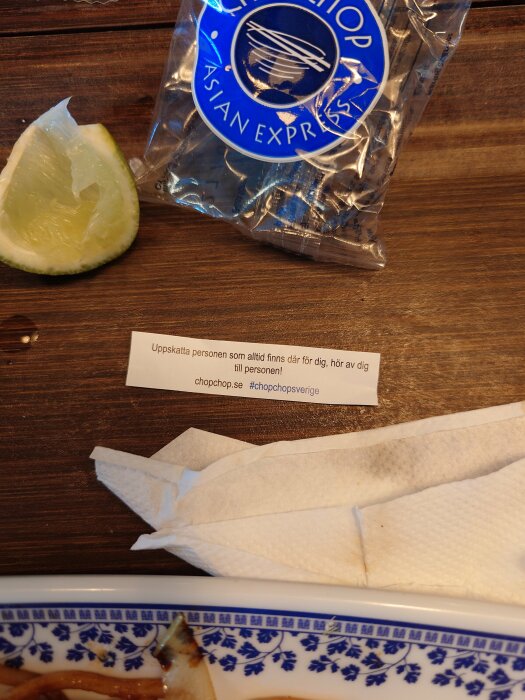 Del av citron, förpackning, pappersservett, meddelande, del av tallrik, matrester, träbord.