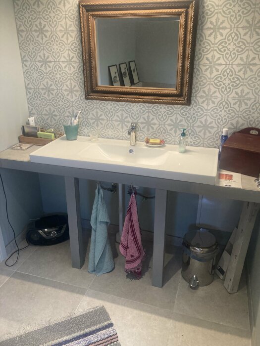 Ett badrum med handfat, spegel, handdukar och personlig hygienartiklar. Mönstrade tapeter och en matta finns också.
