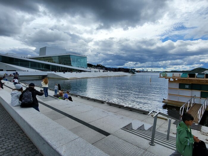 Modern byggnad vid vattnet, molnig himmel, människor vilar och går, stadsmiljö, arkitektoniskt landmärke, brygga, avkoppling.