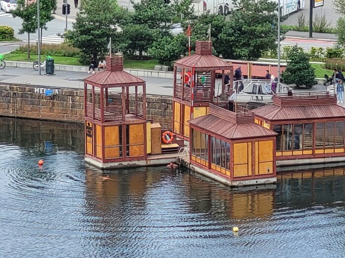 Två flytande byggnader vid kajkanten, människor i bakgrunden, urbant landskap, lugnt vatten, molnig dag.