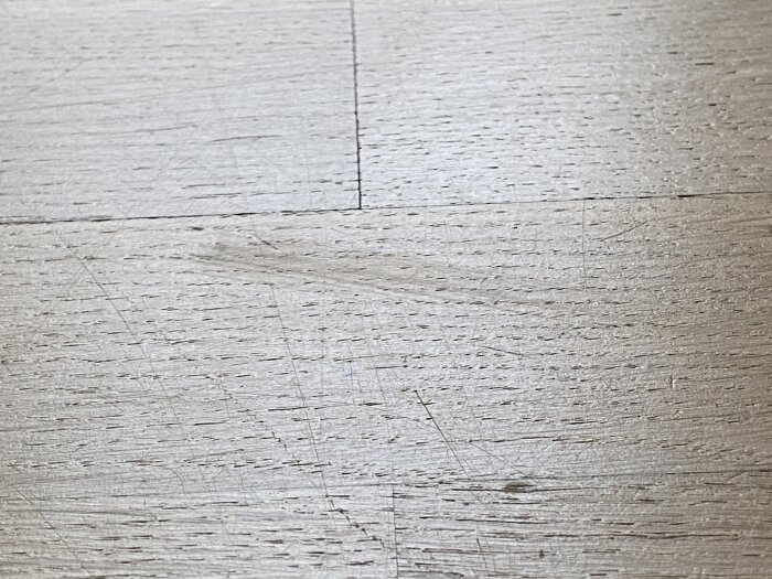 Ett närbildsfoto av texturerat, slitet ljusgrått laminat- eller trägolv med synliga skarvar och repor.