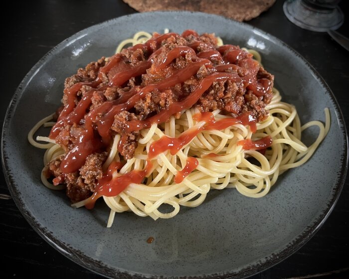 En tallrik med spaghetti och köttfärssås täckt med ketchup, serverad på en mörkgrå keramiktallrik.