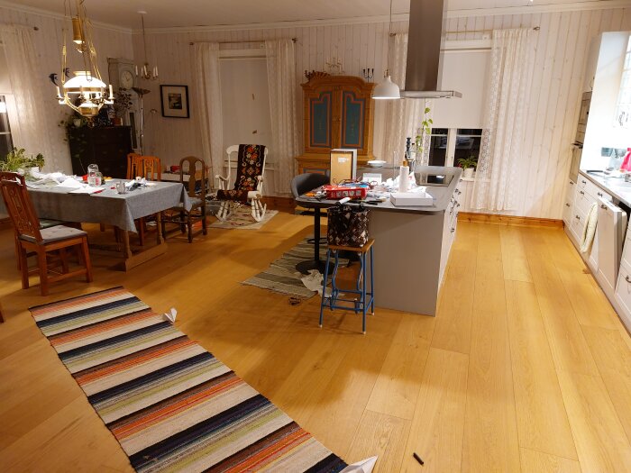 Vardagsrum och kök i öppen planlösning, bord med saker, parkettgolv, mattor, hemtrevlig inredning.