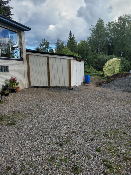 En vit, tegelbyggnad och ett garage med grusuppfart, grönska, molnig himmel, hög med täckt material till höger.