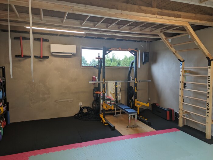 Hemmagym med squat rack, skivstång, bänk, gummigolv, klätterrep och pull-up bars. Träbjälkar i taket, fönster.