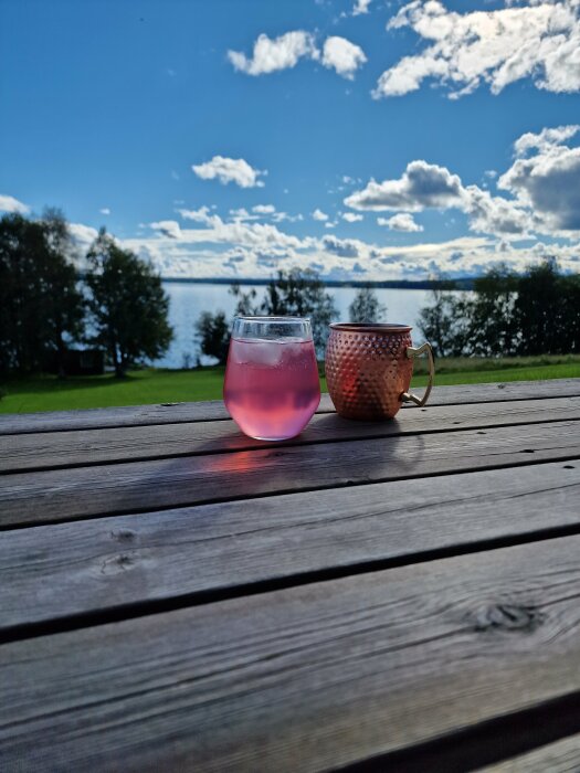 Två drycker på träbord, utsikt över sjö och blå himmel med moln.