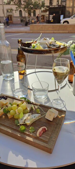 Utomhus, ostbricka, vindruvor, fikon, ett glas vin, östers, ciderflaska, gatumiljö i bakgrunden.