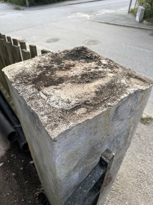 Sliten betongpelare vid vägkant, fläckig yta, närbild, otydlig bakgrund med staket och gata.