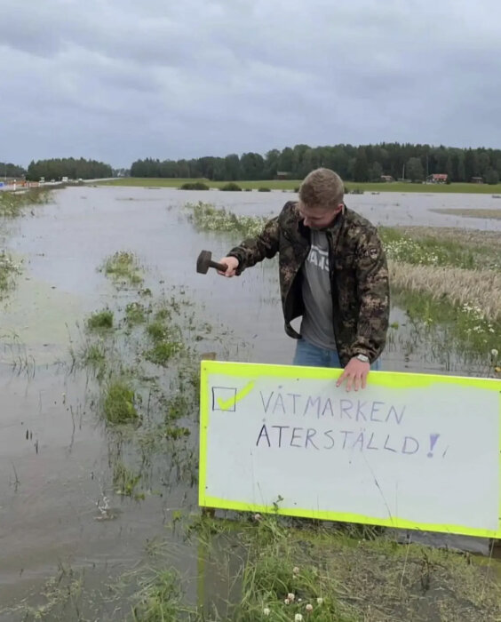 Man med skylt i översvämmat område, "Våtmarken återställd!", humoristiskt budskap.