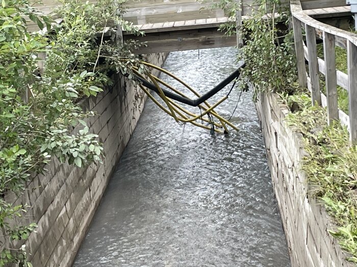 Hammock inlagd ovanför rinnande vatten mellan två träkonstruktioner, omgiven av grönska och räcken.