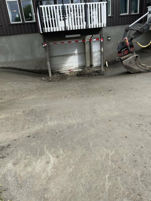 Grävmaskin nära hus, jordhög, skyltade säkerhetsbarriärer under veranda, grått yttervägg, fönster, utomhus.