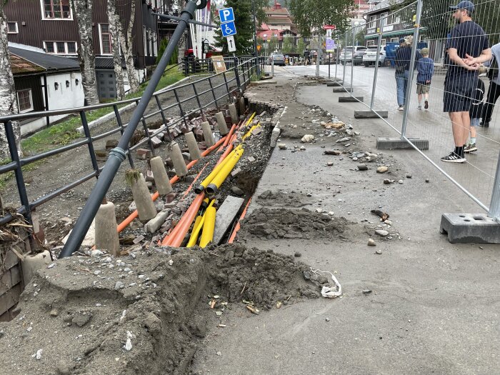 Gatumiljö med grävning, kablar exponerade, skyddsstaket, människor och befintliga byggnader.