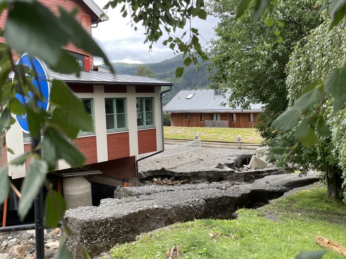 Rödfärgat hus, förstörd asfalt, grönska, bergslandskap i bakgrunden, skylt med blå cirkel, grus och sten.