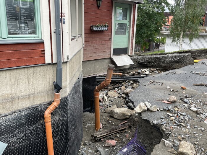 Byggnad med vattenskador, underminerad grund, utsatt för erosion eller jordskred, rör och skräp synliga.